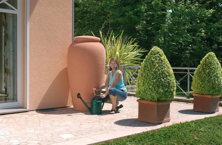 Une femme souriante remplissant un arrosoir à partir d'un grand récupérateur d'eau de pluie en terracotta de 500 litres situé à côté de sa maison, avec de belles plantes en pot et une verdure luxuriante en arrière-plan.