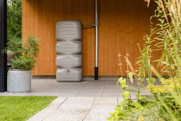 Récupérateur d'eau de pluie vertical de 1000 litres couleur gris béton installé contre un mur en bois, avec un design épuré et un robinet en bas, intégré dans un jardin verdoyant.