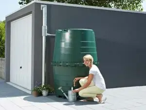 Une femme souriante utilise un robinet de récupérateur d'eau de pluie Garantia de 1000L dans un jardin moderne, démontrant une utilisation pratique et écologique de la récupération d'eau.